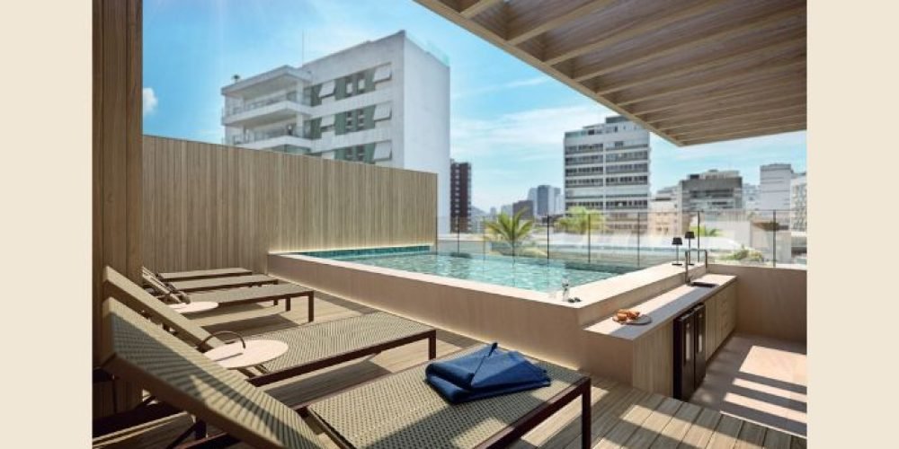 Guilhem Mozak Leblon - Espaço de uso comum com mobiliário em madeira clara, mesas e cadeiras de design moderno, em ambiente iluminado naturalmente por janelas amplas.